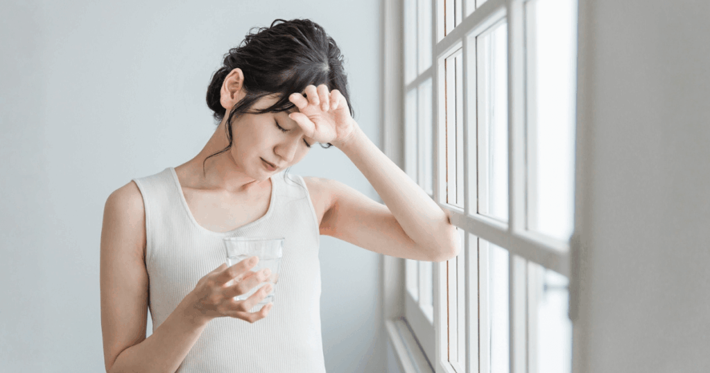 体調不良で不安そうに額へ手を当てながら水を持つ女性の写真