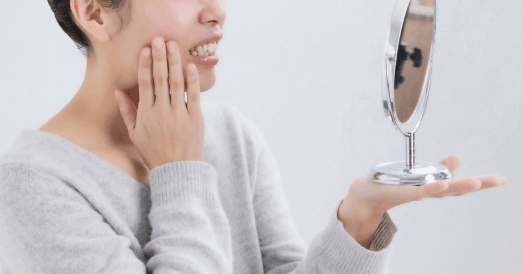 鏡を見ながら口元の違和感を気にして頬に手を当てる女性の様子